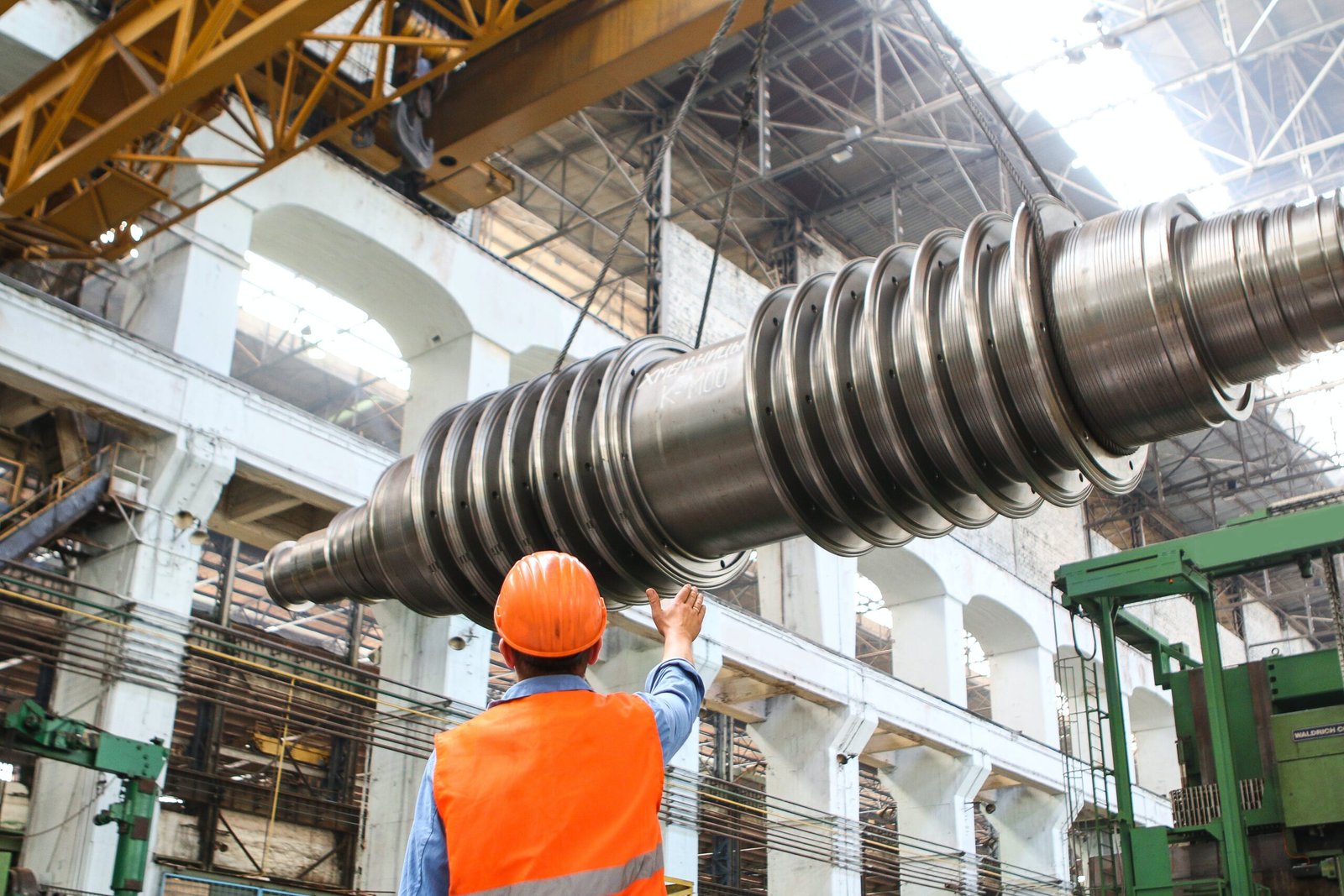 a labour project manager pointing at a crane which holds a big part in a manufacturing factory